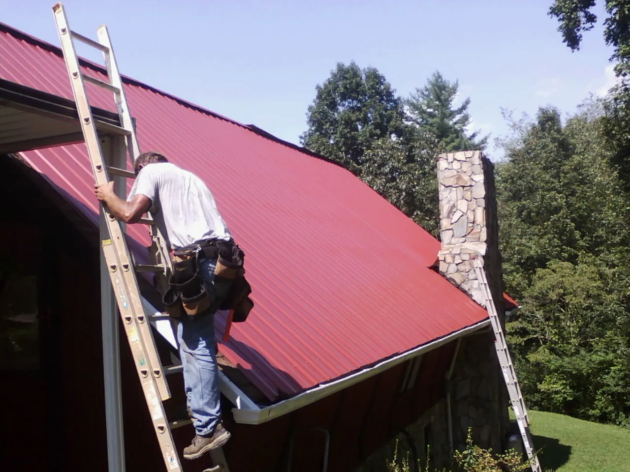 Roofer on a ladder beside a red metal roof and stone chimney — Soffit & Fascia Repair by Roofing Brothers in Greenfield